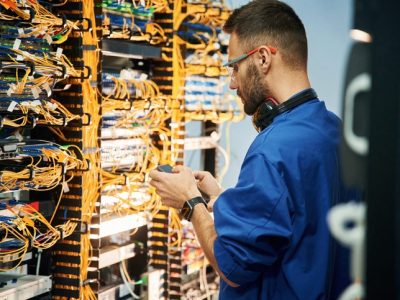 Holding smartphone. Young man is working with internet equipment and wires in server room
